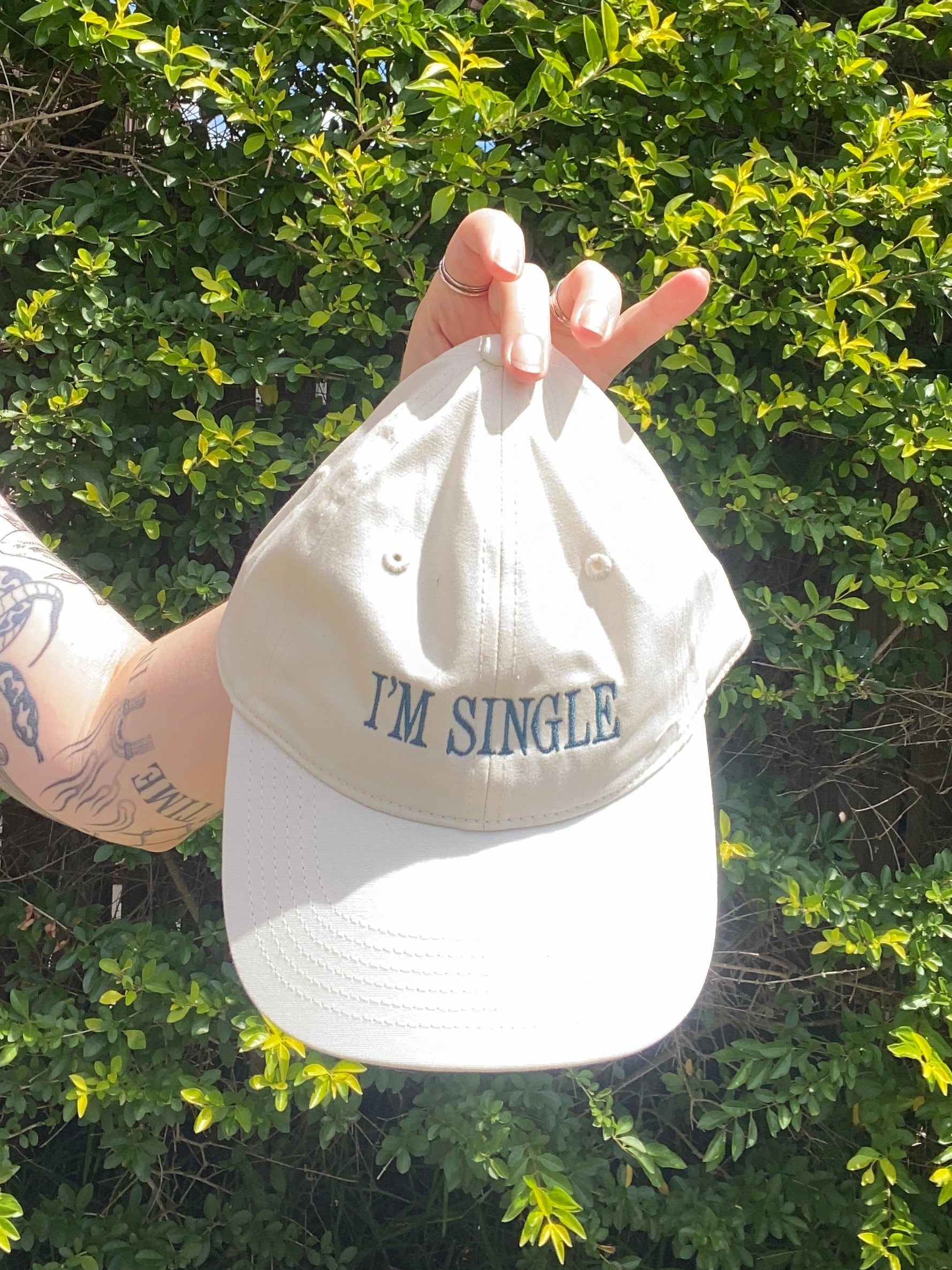 Hand holding a beige cap with text against a green bush background